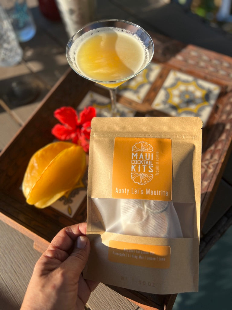 Hand holding a tropical cocktail kit package with a cocktail and ingredients on a table.