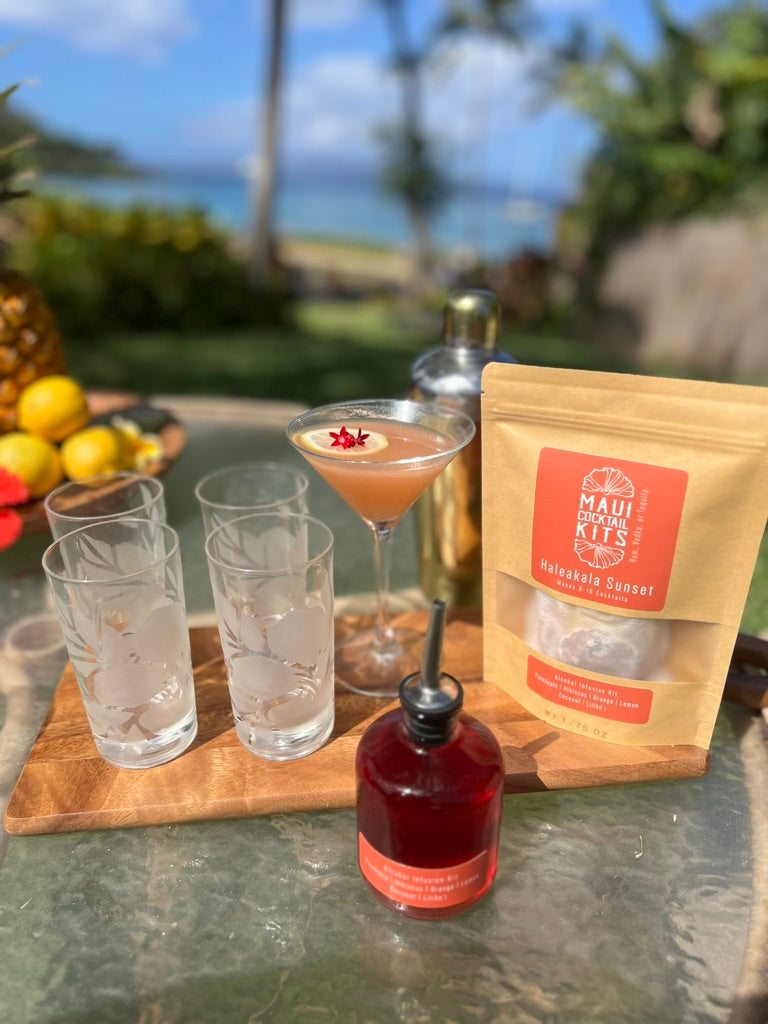 Tropical cocktail setup with glasses, bottle, and Maui Cocktail Kits bag on a table outdoors.